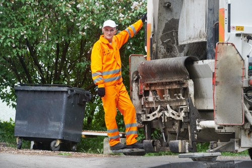 A municipal waste collection worker dressed in high-visibility orange protective clothing, including a white cap, stands beside a large urban rubbish collection vehicle. The worker is positioned on a paved street near green foliage, with one hand resting on the vehicle as they observe or prepare for waste collection. The vehicle features a large metal compactor and a chute for loading rubbish, with reflective safety markings along the sides. Nearby, a substantial black wheeled bin is placed on the pavement, ready for disposal, indicating an outdoor rubbish removal service. The background includes trees with dense green leaves, contributing to an outdoor, residential or urban environment. The scene is lit naturally, highlighting the details of the worker’s attire, the textures of the bin and vehicle, and the surrounding greenery, reflecting typical rubbish collection procedures that a company such as Garden Clearance Ilford might perform in the Ilford area.