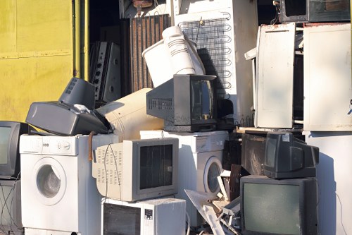 A large collection of discarded household appliances and electronic waste is piled in an outdoor storage area, including washing machines, microwave ovens, and computer monitors with visible screens. The appliances are made of metal, plastic, and glass, with some showing signs of dirt and weathering. They are arranged haphazardly, with items leaning against each other and some partially covered by shadows cast by a nearby structure. In the background, a corrugated metal wall and a yellow panel are visible, indicating an industrial or waste management site. The overall scene depicts a typical rubbish collection site in Ilford, where Garden Clearance Ilford manages outdoor waste removal services, emphasizing the disposal of bulky household items. Natural light highlights the textures of the materials, underscoring the need for professional rubbish clearance to properly handle and remove this type of waste from residential or commercial properties in the local area.